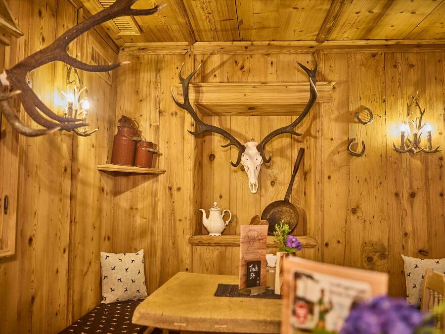 A rustic wooden interior with a table, chairs, a shelf with jars, a skull, and wall-mounted lights.