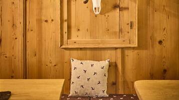 Interior de madera con un cráneo de ciervo montado en la pared, un banco de madera con cojines estampados y una mesa con una lámpara.