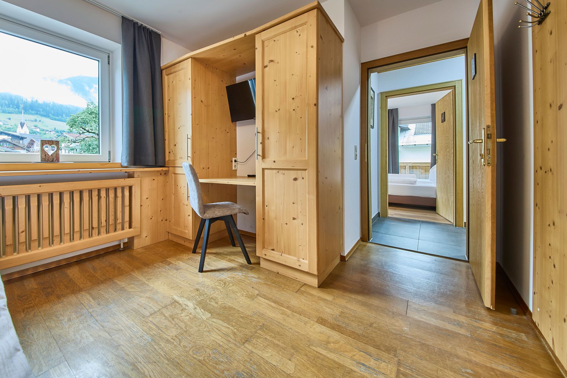 A room with wooden flooring, a desk with a chair, a TV mounted on the wall, and a bed in the adjacent room.