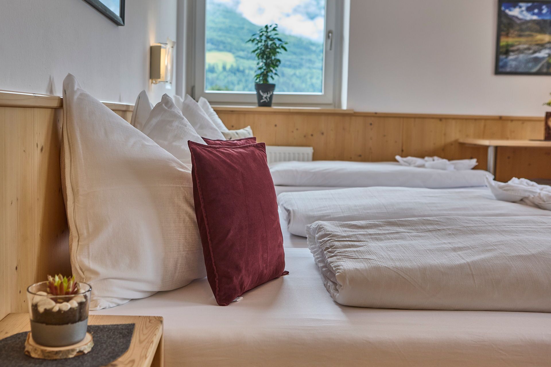 A bedroom with two beds, white bedding, a red pillow, a wooden headboard, a bedside table with a glass container, and a window with a view of greenery.