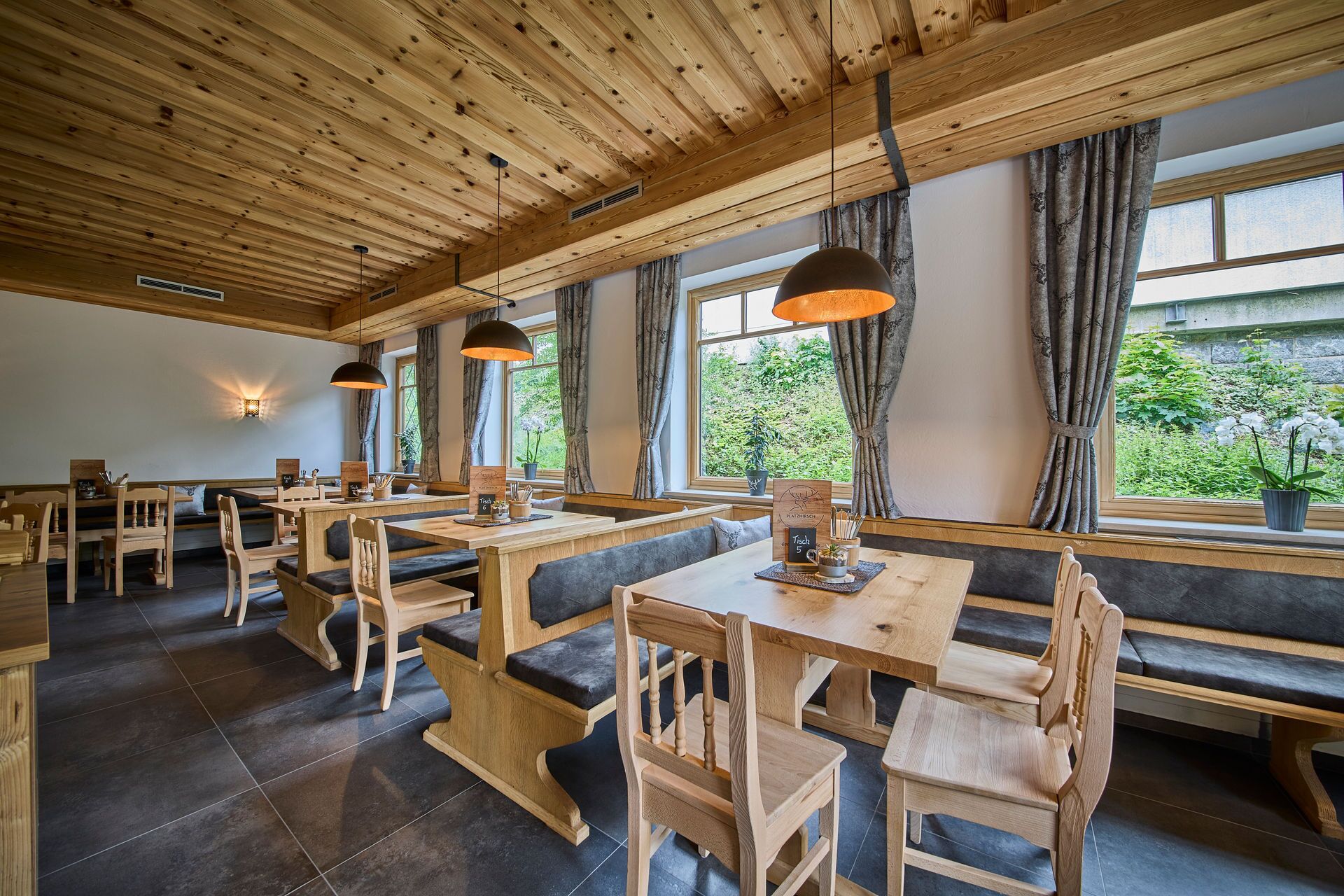 A restaurant with wooden tables and benches, large windows with curtains, and pendant lighting.