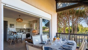 An outdoor dining area with a table set for four, a kitchen in the background, and a view of trees and a clear sky. - Les Marines de Gassin by Interhome (Gassin)