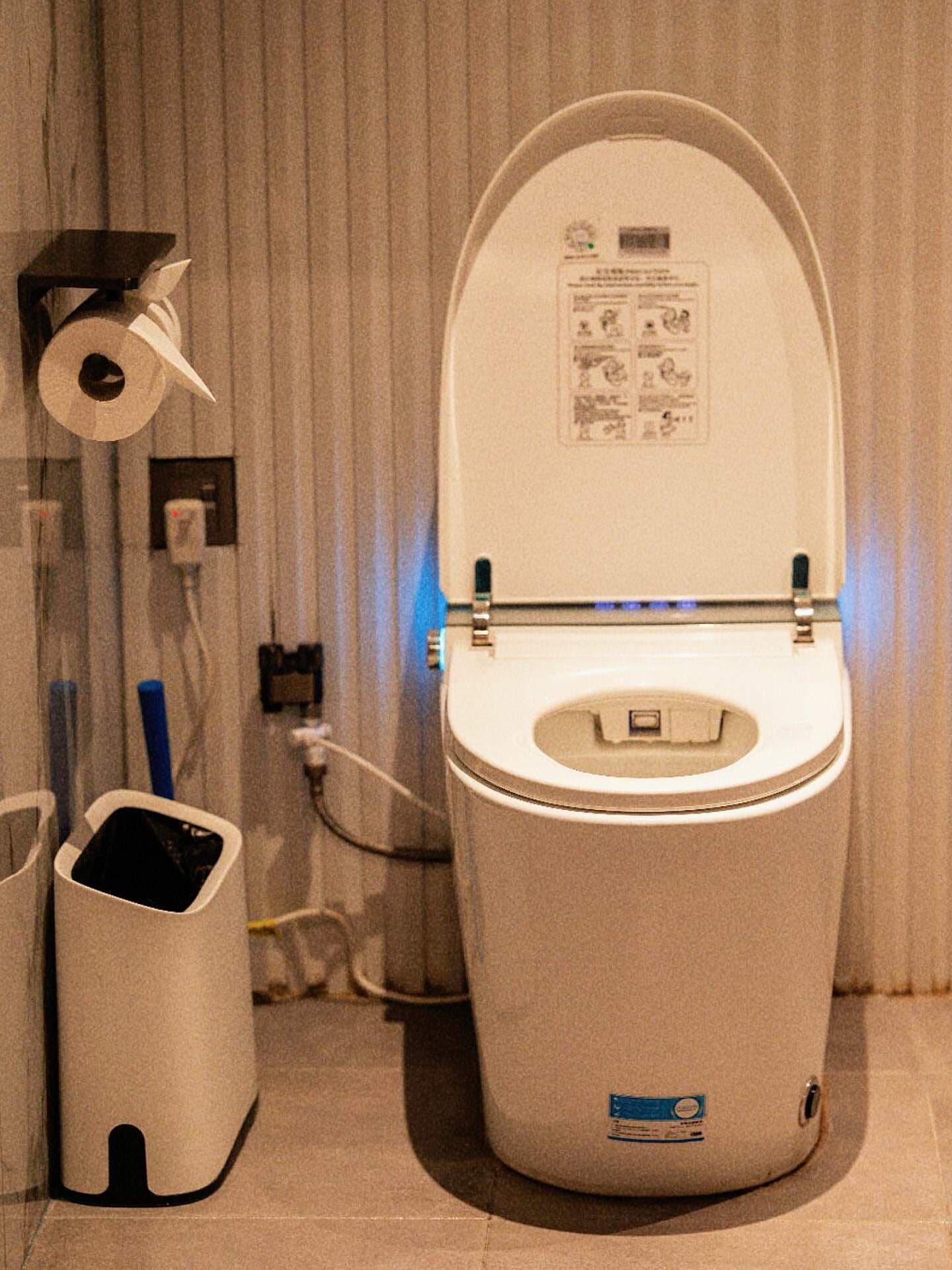 Des toilettes blanches avec un abattant fermé, un porte-rouleau de papier toilette et une poubelle dans une salle de bain avec un rideau à rayures.