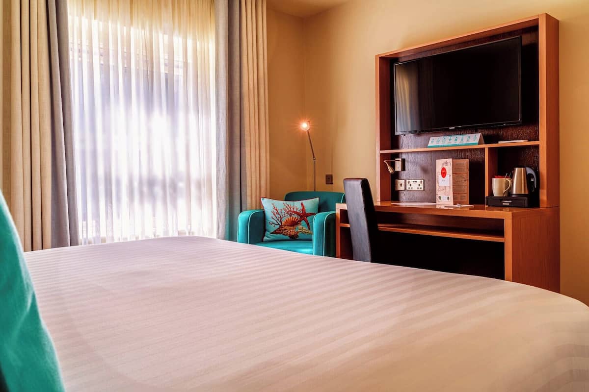 A hotel room with a bed, a desk with a flat-screen TV, a coffee machine, and a chair with a decorative pillow.