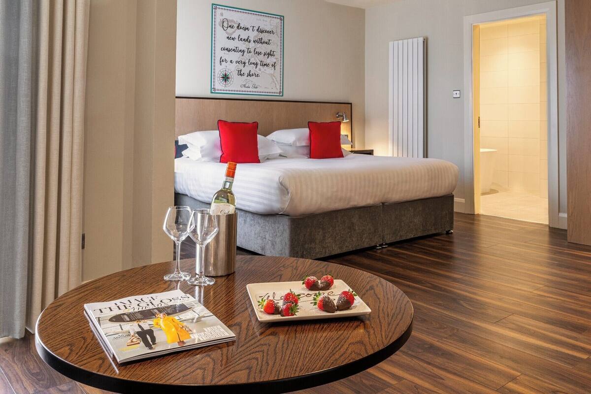 A hotel room with a bed, a bedside table with a bottle of wine and glasses, a magazine, and a plate of strawberries.