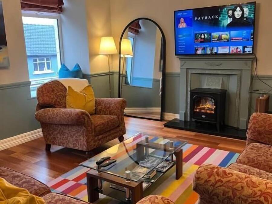 Una sala de estar con sofás estampados, una mesa de centro de vidrio, una chimenea y un televisor mostrando una película.