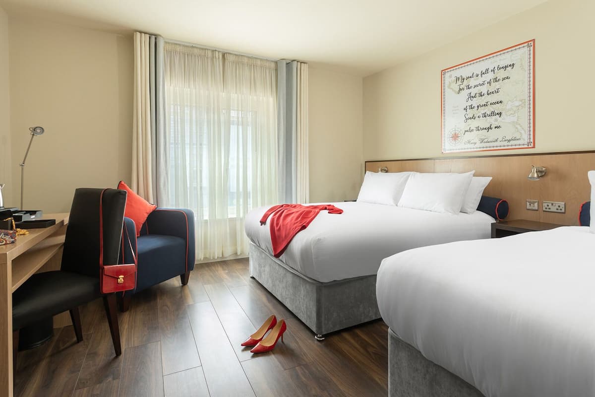 A hotel room with two beds, a desk with a chair, a lamp, and a large window with curtains.