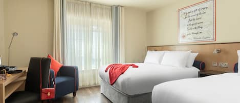 Habitación de hotel con dos camas, un escritorio con silla, una lámpara y un ventanal con cortinas.