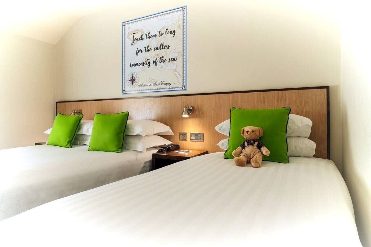 A hotel room with two beds, green pillows, a wall-mounted sign, and a teddy bear on one bed.