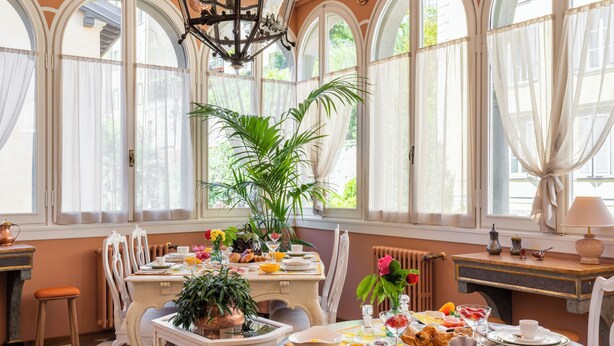 A dining area with a table set for breakfast, surrounded by large windows with sheer curtains, a chandelier, and a radiator. - Palazzo Radici (Bergamo)