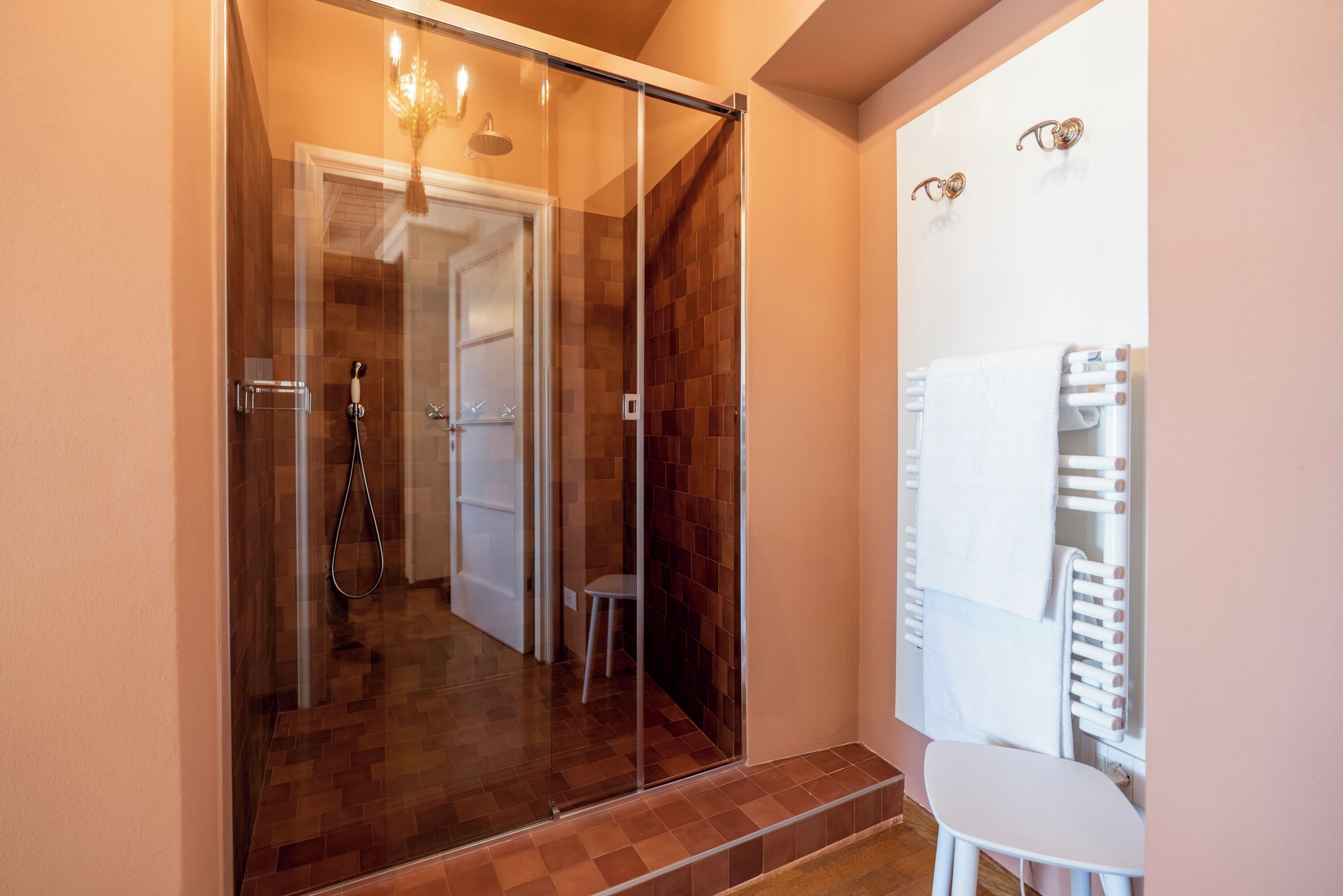 Área de chuveiro com paredes de vidro, uma cadeira branca, porta-toalhas e uma porta visível que dá para outro cômodo.