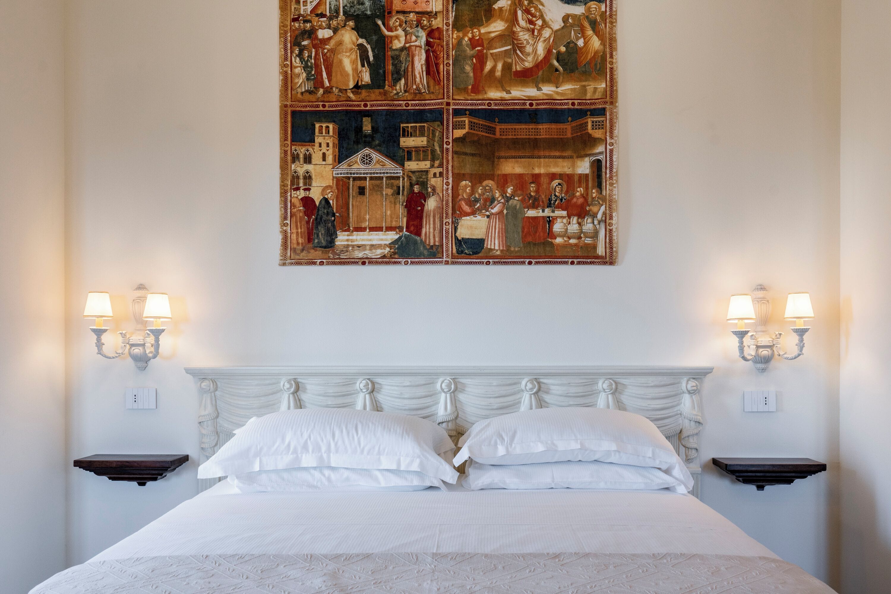 A neatly made bed with white linens and two bedside tables with wall-mounted lamps.