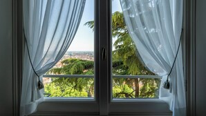 A window with white curtains and a view of greenery and buildings. - Palazzo Radici (Bergamo)
