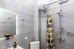 A bathroom with grey tiles, a toilet, a sink with a mirror, a shower with a rain showerhead, and a patterned shower curtain. - Anya Studios by Estia (Corfu)