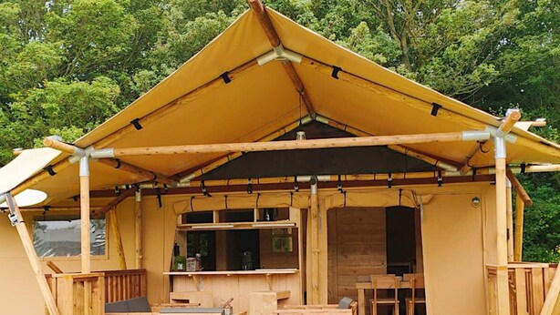 A wooden cabin with a covered porch, built-in seating, and a small kitchen area. - Nice Tent With Bathroom, Near a Recreation Area (Noord-Scharwoude)