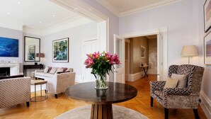 A spacious living room with a round wooden table, a white sofa, and a patterned armchair. - Majestic in Kensington, by Veeve (London)
