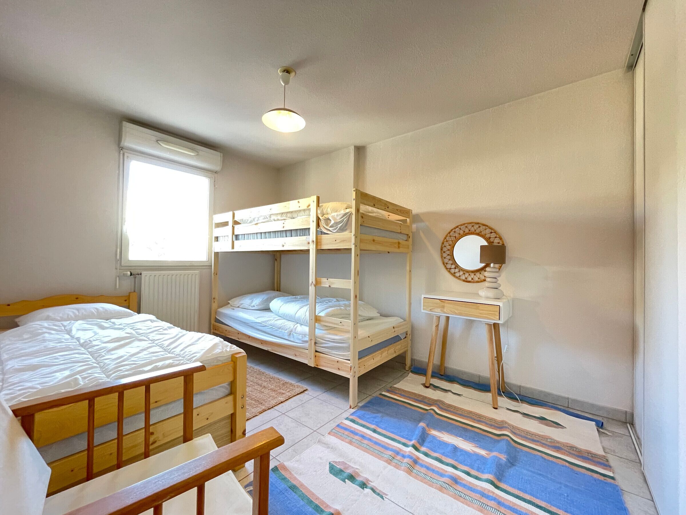 A room with bunk beds, a bedside table with a lamp, a mirror, and a window with blinds.