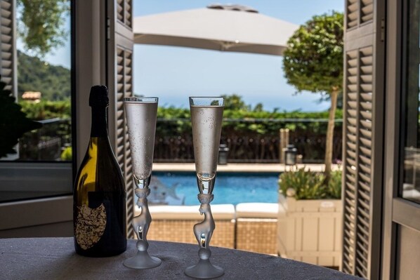 A bottle of champagne and two champagne flutes on a table with a view of a swimming pool and greenery.