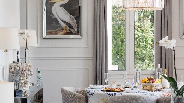 Un salon moderne avec un canapé gris, un lustre, une œuvre d’art représentant une mouette encadrée et une table à manger dressée pour un repas.