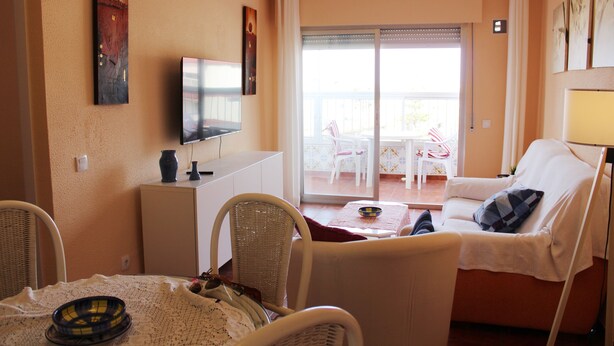 A living room with a television, wicker chairs, a dining table with a bowl, and a balcony with a table and chairs. - M & M Neptuno Torrecilla Beach. With Pool (Nerja)