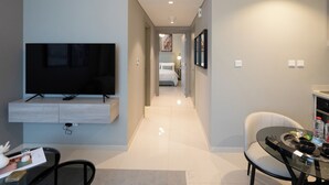A modern hotel room with a flat-screen TV mounted on a wooden panel, a dining area with a glass table and a white chair, and a hallway leading to a bedroom. - Business Bay Bliss 1BR Apartment (Dubai)