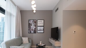A modern living room with a grey sofa, a small round table, and a flat-screen TV on a stand. There are two framed pictures on the wall and a window with sheer curtains. - Business Bay Bliss 1BR Apartment (Dubai)