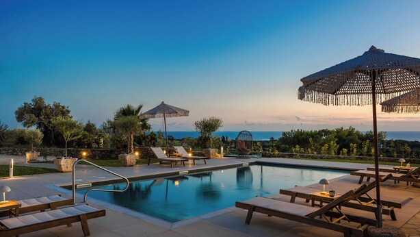Zona de piscina con sillones reclinables, sombrillas y una vista despejada del océano al atardecer.