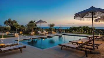 Un espace aménagé au bord de la piscine, avec des chaises longues, des parasols et une vue dégagée sur l’océan au crépuscule.