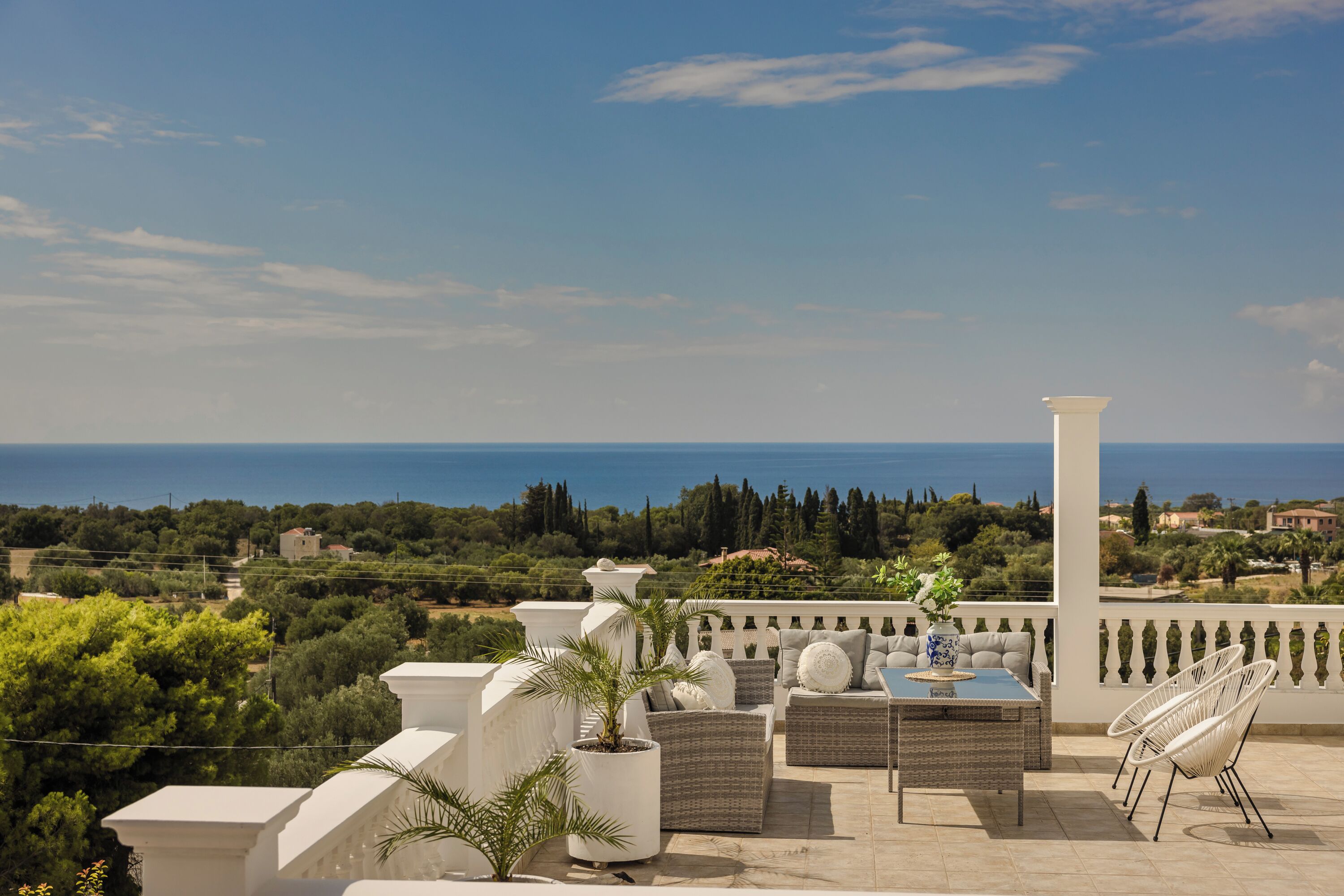 Une terrasse aménagée avec du mobilier d’extérieur, une colonne blanche et une vue sur la mer et la verdure.