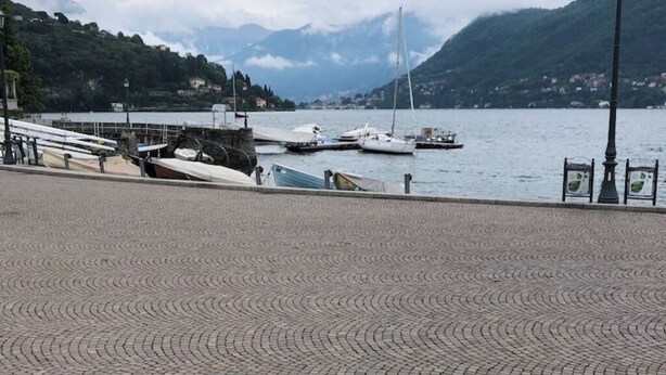 Une promenade en bord de mer, dallée, avec des bateaux amarrés, un mur de pierre et une chaîne de montagnes en arrière-plan.