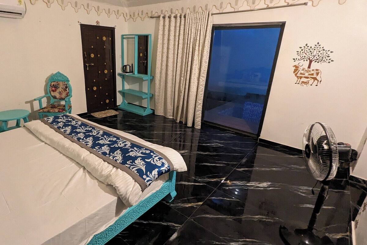 A hotel room with a bed, a small table, a chair, a fan, and a view of the outside.