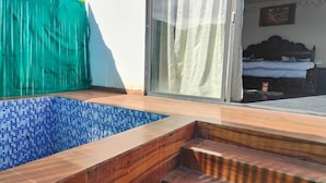 Une piscine aux carreaux de mosaïque bleus, des marches en bois menant à une porte, et un lit avec une tête de lit sombre en arrière-plan.