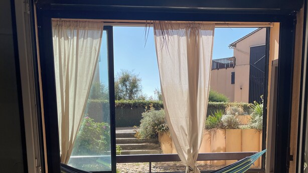 A room with a large window, a hammock, and a view of a courtyard with plants and a building in the background.