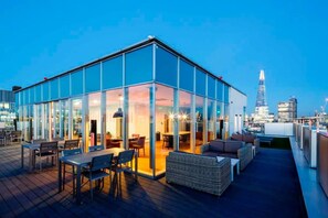 Um edifício moderno de vidro com terraço no telhado, mobiliário para área externa e vista para o horizonte da cidade.