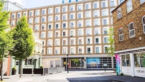 A modern building with a grid of windows, a storefront with a colorful sign, and a tree in the foreground. - For Students Only Chapter South Bank in London (London)