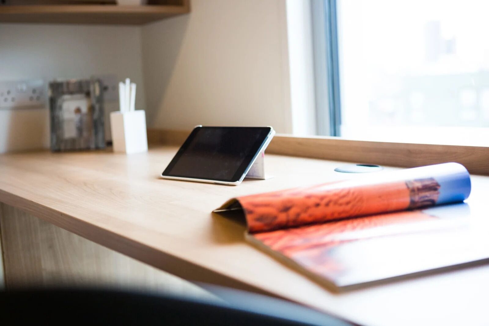 A wooden desk with a tablet, a rolled-up magazine, and a pen holder.