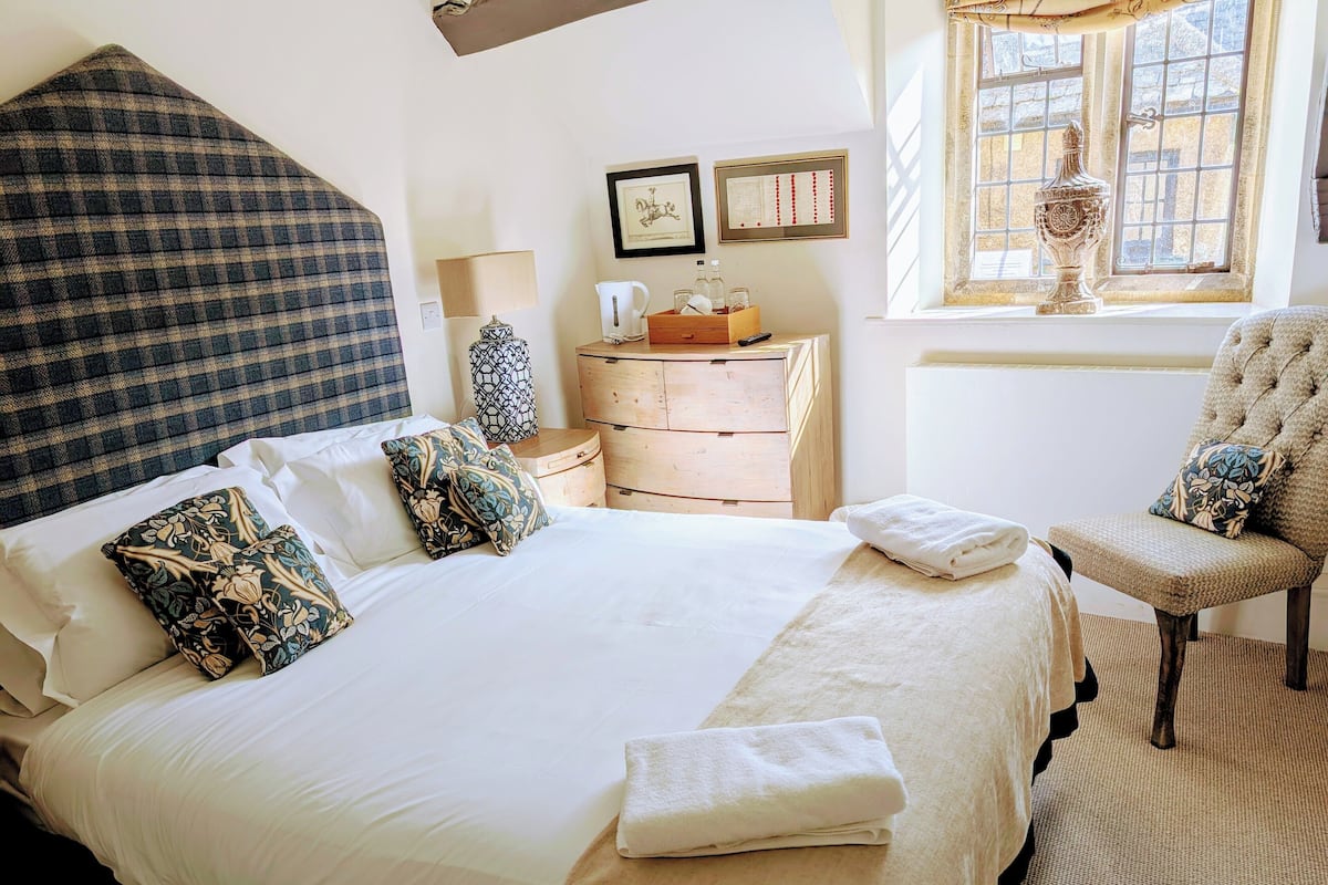 A neatly made bed with white linens and decorative pillows, a wooden nightstand with a lamp, a dresser, and a chair with a cushion.