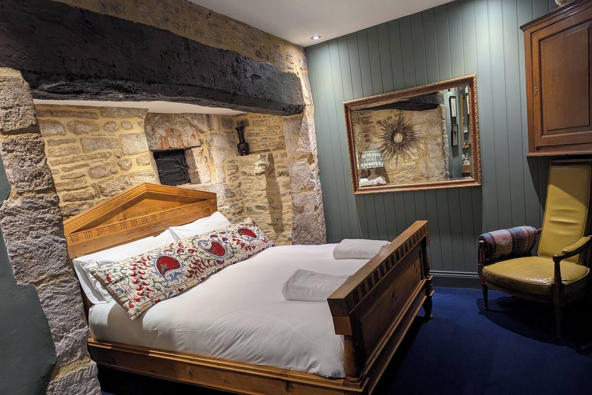 A bedroom with a stone wall, a wooden bed, a mirror, and a chair.