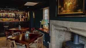 A bar area with a variety of liquor bottles, a wooden dining table with chairs, a fireplace with a stone mantle, and a painting on the wall. - The Crown at Shipton (Chipping Norton)