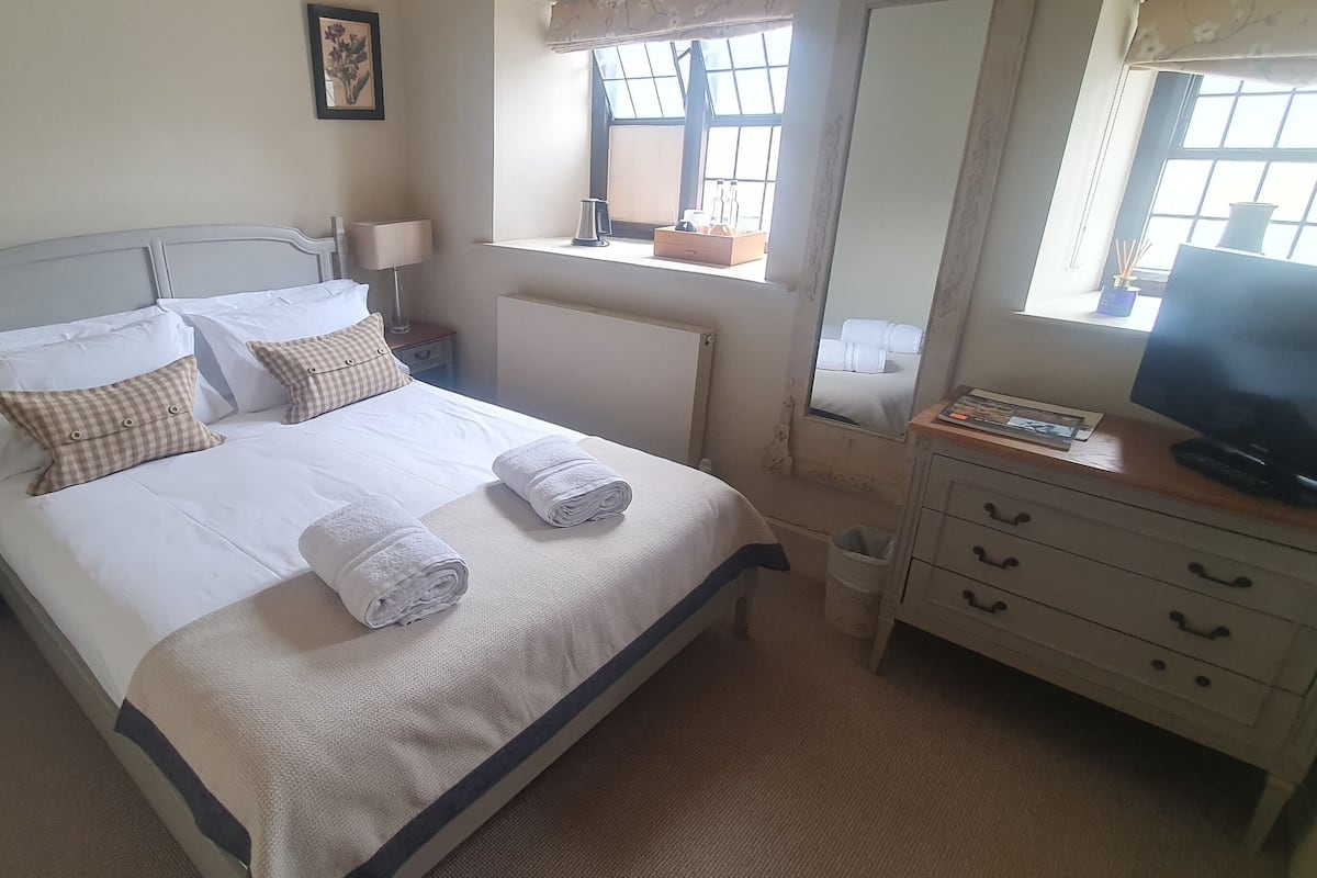 A neatly made bed with pillows and a rolled towel, a bedside table with a lamp, a dresser with a mirror, and a TV on a wooden stand.