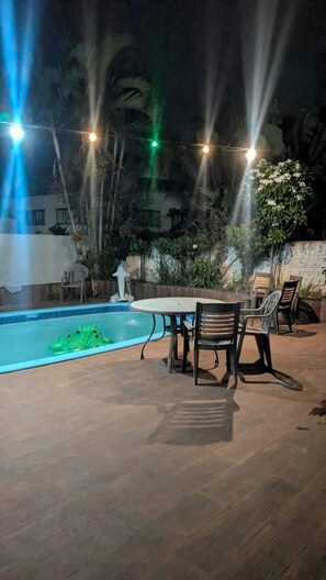 Zona de piscina con mesa y sillas, rodeada de plantas y un muro de piedra.