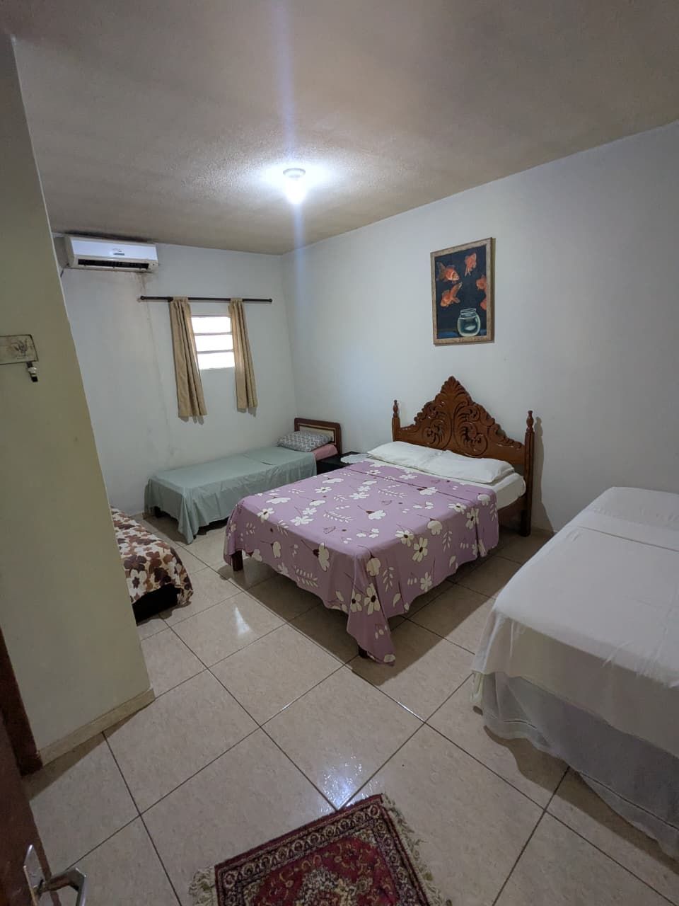 Ein Schlafzimmer mit zwei Betten, einem Deckenventilator, einer Klimaanlage, einem Fenster mit Vorhängen und einem Bild an der Wand.