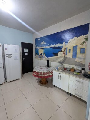 Una cocina con suelo de baldosas, un refrigerador, una cafetera, una mesa con mantel estampado de flores y un mural en la pared que representa una escena costera.