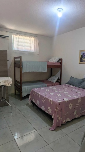 Un dormitorio con literas, una cama individual con colcha estampada, una mesita con mantel de encaje y una ventana con cortinas.
