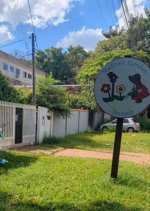 A sign for 'Meliza's Garden' with a birdhouse and flowers, a white fence, and a parked car. - Melizas Garden (Foz do Iguaçu)