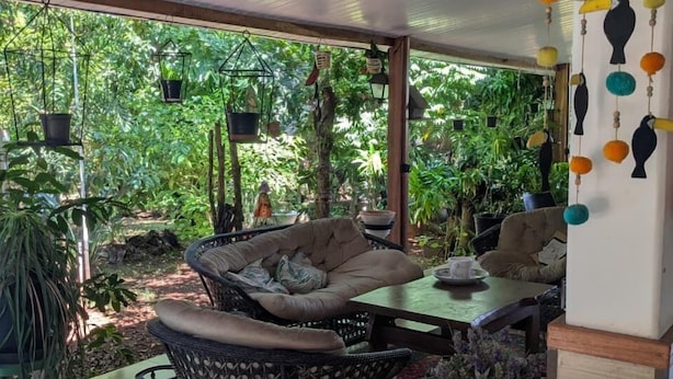 Eine überdachte Terrasse mit Korbmöbeln, einem Tisch, hängenden Pflanzen und einer dekorativen Vogelstatue.