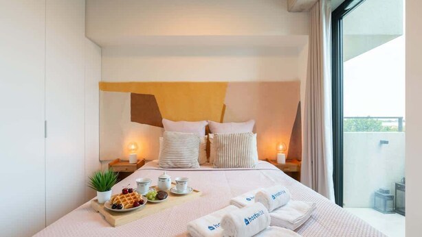 Een moderne hotelkamer met een groot bed, een nachtkastje met lamp, een dienblad met wafels en fruit, en uitzicht op de omgeving.