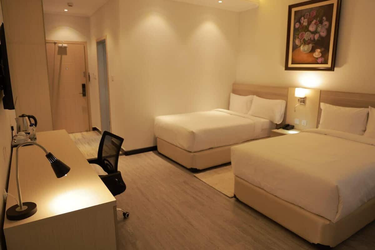 A hotel room with two beds, a desk, and a chair. There is a painting on the wall and a door in the background.