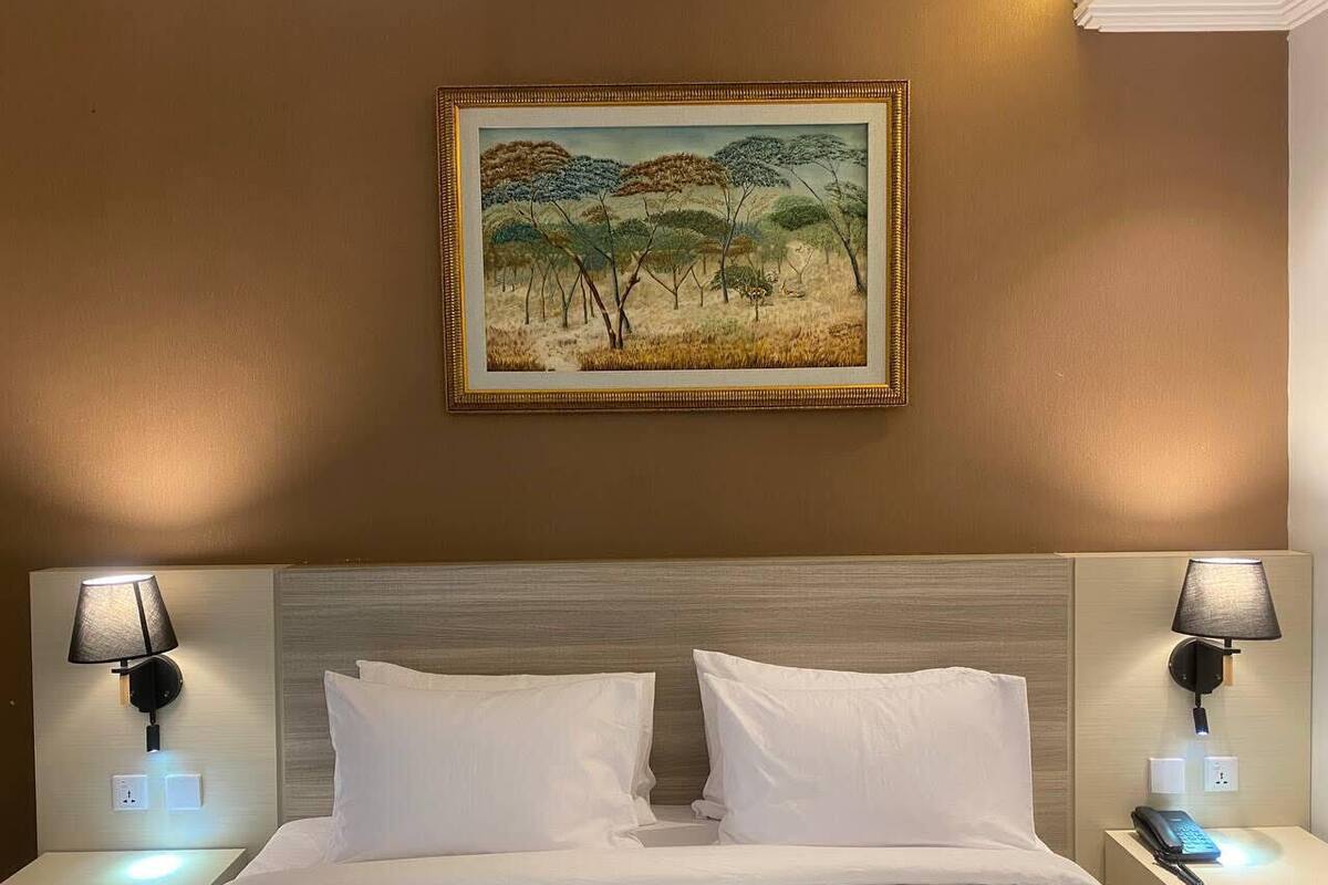 A hotel room with a bed, two bedside tables, a painting on the wall, and a nightstand with a lamp.