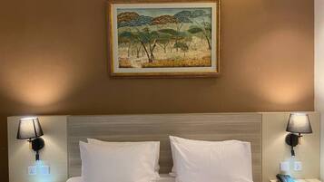 A hotel room with a bed, two bedside tables, a painting on the wall, and a nightstand with a lamp.